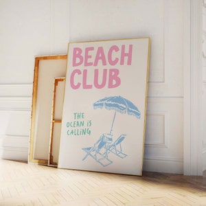 Pu&ograve; includere: Un poster bianco con cornice dorata che presenta il testo "Beach Club" in lettere rosa e il testo "The ocean is calling" in lettere verdi. Un ombrellone da spiaggia blu e due sedie a sdraio blu sono illustrati sotto il testo.