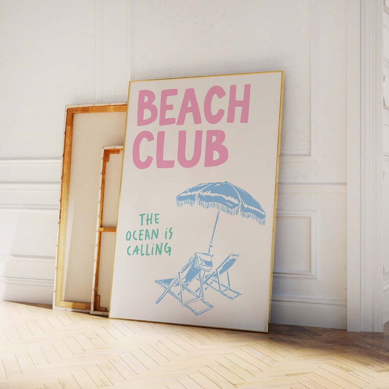 Pu&ograve; includere: Un poster bianco con cornice dorata che presenta il testo "Beach Club" in lettere rosa e il testo "The ocean is calling" in lettere verdi. Un ombrellone da spiaggia blu e due sedie a sdraio blu sono illustrati sotto il testo.
