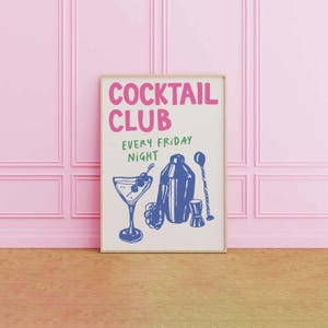 May include: A framed print with the words "COCKTAIL CLUB EVERY FRIDAY NIGHT" in pink and green. The print features blue illustrations of a cocktail glass, shaker, and bar tools. The background is a light cream color.