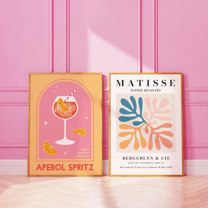 May include: Two framed art prints. The print on the left is a pink and orange illustration of an Aperol Spritz cocktail with the text "Aperol Spritz" and the ingredients listed below. The print on the right is a colorful abstract art print with the text "Matisse Papier Decoup&eacute;s" and "Berggruen & Cie" with the date "Du Vendredi 27 Fevrier au Samedi 28 Mars 1953".