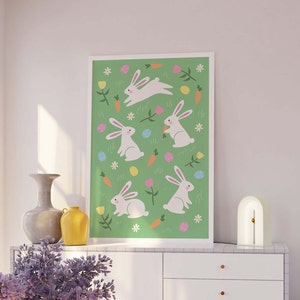 May include: A green print with white bunnies, carrots, and colorful eggs. The print is framed and hanging on a white wall above a white dresser.