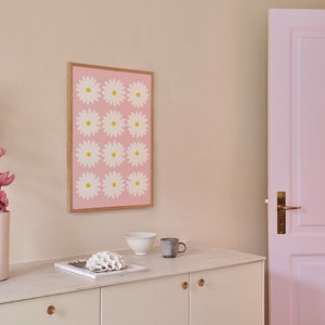 May include: A white cabinet with two doors and gold knobs. The cabinet is sitting on a white countertop with a white bowl, a brown mug, and a white coral sculpture on top. A framed print with a pink background and white flowers with yellow centers is hanging on the wall behind the cabinet. A pink door is visible on the right side of the image.