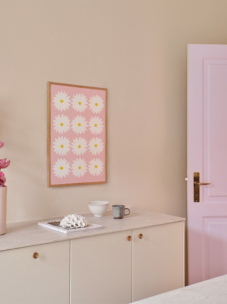 May include: A white cabinet with two doors and gold knobs. The cabinet is sitting on a white countertop with a white bowl, a brown mug, and a white coral sculpture on top. A framed print with a pink background and white flowers with yellow centers is hanging on the wall behind the cabinet. A pink door is visible on the right side of the image.