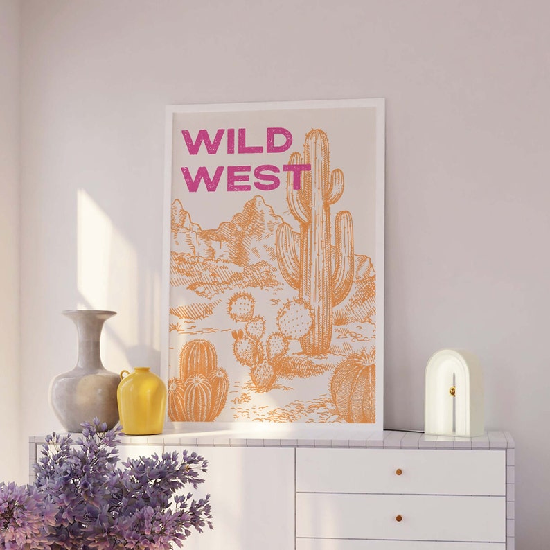 May include: A white framed print with the text "WILD WEST" in pink lettering. The print features a line drawing of a desert landscape with a large cactus in the center.