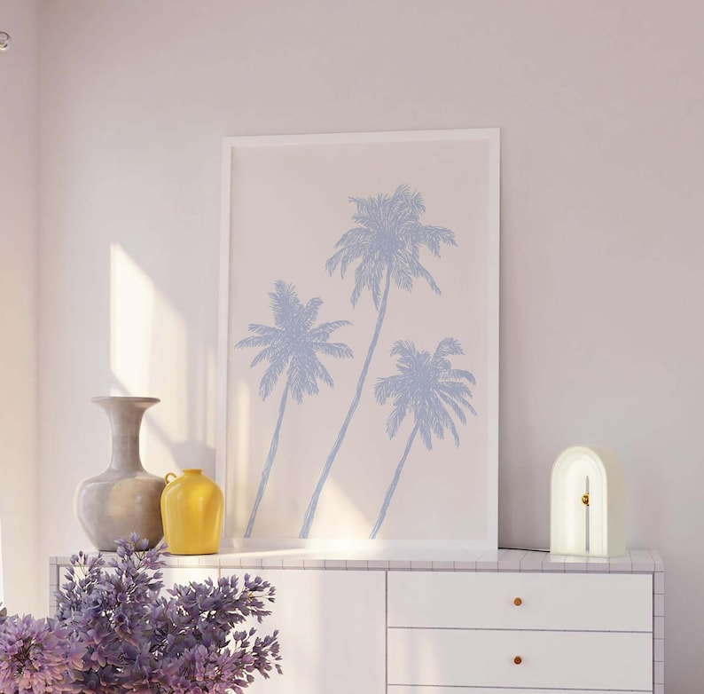 Pode incluir: Uma impress&atilde;o emoldurada branca com uma ilustra&ccedil;&atilde;o azul claro de tr&ecirc;s palmeiras sobre um fundo branco. A impress&atilde;o est&aacute; sobre uma c&ocirc;moda branca com duas gavetas. Um vaso cinza e um vaso amarelo est&atilde;o sobre a c&ocirc;moda. Uma l&acirc;mpada branca em forma de arco com um acento dourado est&aacute; no lado direito da c&ocirc;moda.