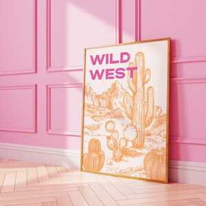 May include: A framed print with the text "WILD WEST" in pink lettering. The print features a hand-drawn illustration of a desert landscape with cacti in orange.