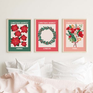 May include: Three Christmas market prints with a green, red, and pink background. The Vienna print features red poinsettia flowers. The London print features a green wreath with red berries. The Paris print features a bouquet of red flowers with green leaves.