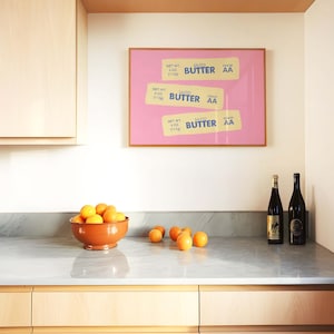 Peut inclure: Int&eacute;rieur de cuisine avec une impression encadr&eacute;e rose de trois &eacute;tiquettes de beurre, un bol en terre cuite rempli d'oranges et deux bouteilles de vin. Les &eacute;tiquettes indiquent "SALTED BUTTER AA" et "113g".