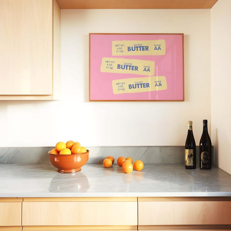 Peut inclure: Int&eacute;rieur de cuisine avec une impression encadr&eacute;e rose de trois &eacute;tiquettes de beurre, un bol en terre cuite rempli d'oranges et deux bouteilles de vin. Les &eacute;tiquettes indiquent "SALTED BUTTER AA" et "113g".