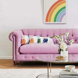 Peut inclure: Un canap&eacute; rose capitonn&eacute; avec une &oelig;uvre d'art encadr&eacute;e repr&eacute;sentant un arc-en-ciel au-dessus. Le canap&eacute; a deux coussins avec un motif g&eacute;om&eacute;trique color&eacute;. Une table basse en verre et en marbre se trouve devant le canap&eacute; avec un vase de fleurs blanches et violettes sur le dessus.