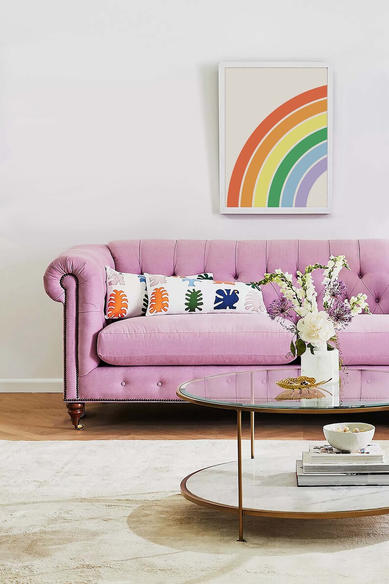 Peut inclure: Un canap&eacute; rose capitonn&eacute; avec une &oelig;uvre d'art encadr&eacute;e repr&eacute;sentant un arc-en-ciel au-dessus. Le canap&eacute; a deux coussins avec un motif g&eacute;om&eacute;trique color&eacute;. Une table basse en verre et en marbre se trouve devant le canap&eacute; avec un vase de fleurs blanches et violettes sur le dessus.