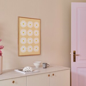 May include: A white cabinet with two doors and gold knobs. The cabinet is topped with a white countertop with a white bowl, a brown mug, and a white coral-like object on top of a magazine. A framed print with a yellow background and white flowers is hanging on the wall behind the cabinet. A pink door is visible on the right side of the image.
