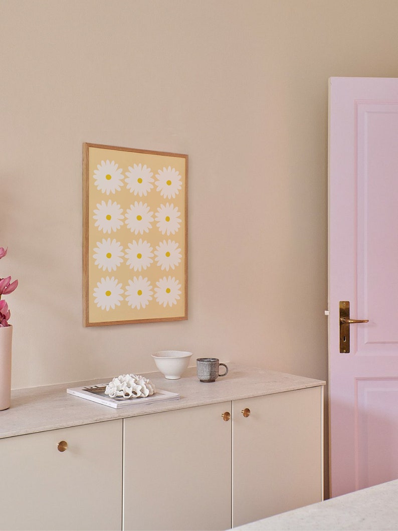 May include: A white cabinet with two doors and gold knobs. The cabinet is topped with a white countertop with a white bowl, a brown mug, and a white coral-like object on top of a magazine. A framed print with a yellow background and white flowers is hanging on the wall behind the cabinet. A pink door is visible on the right side of the image.