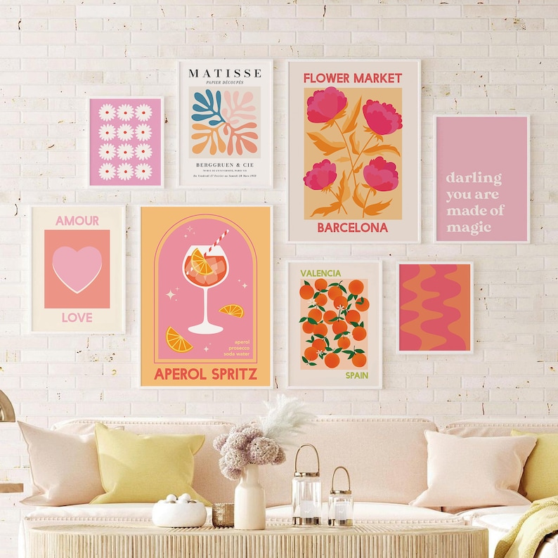 May include: A set of nine colorful wall art prints with various designs, including a heart, flowers, oranges, and a cocktail. The prints are framed in white and have pink, orange, and yellow accents.