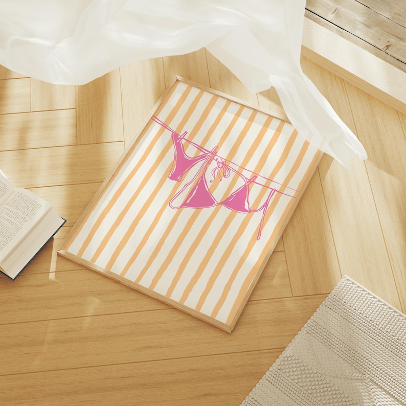 May include: A framed print featuring a pink bikini hanging on a clothesline against a backdrop of orange and white stripes. The artwork is set on a wooden floor, with a book and a white curtain in the background. The overall theme is summer and beach.