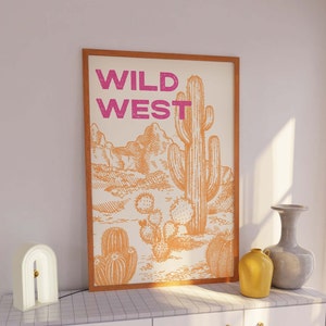 May include: A framed print with the text "WILD WEST" in pink lettering on a white background. The print features a hand-drawn illustration of a desert landscape with a large cactus in the center.