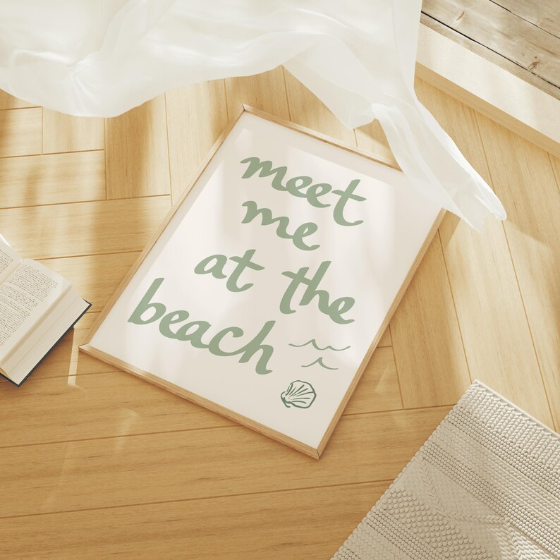 Puede incluir: Un cuadro enmarcado con las palabras "meet me at the beach" en escritura verde claro. La impresi&oacute;n incluye ilustraciones de olas y conchas marinas. El marco es de madera clara y el fondo es de color crema. La impresi&oacute;n est&aacute; sobre un suelo de madera.