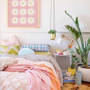 May include: A pink and white bedroom with a bed, a white nightstand, a hanging plant, and a framed print of white daisies on a pink background.