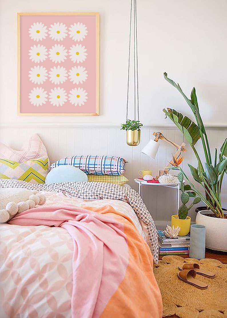 May include: A pink and white bedroom with a bed, a white nightstand, a hanging plant, and a framed print of white daisies on a pink background.