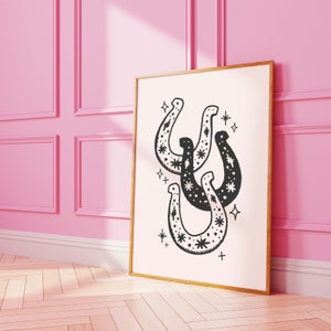 May include: A black and white print of three horseshoes with stars inside them. The print is framed in a gold frame and is hanging on a pink wall.