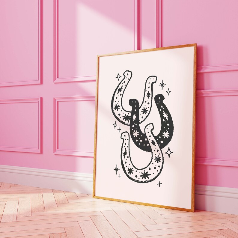 May include: A black and white print of three horseshoes with stars inside them. The print is framed in a gold frame and is hanging on a pink wall.