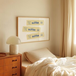 May include: A framed print of three vintage-style butter labels with the text "SALTED BUTTER GRADE AA" and the net weight of 4 oz (113g). The print is hanging on a wall in a bedroom with a white lamp and a bed with a beige linen duvet.