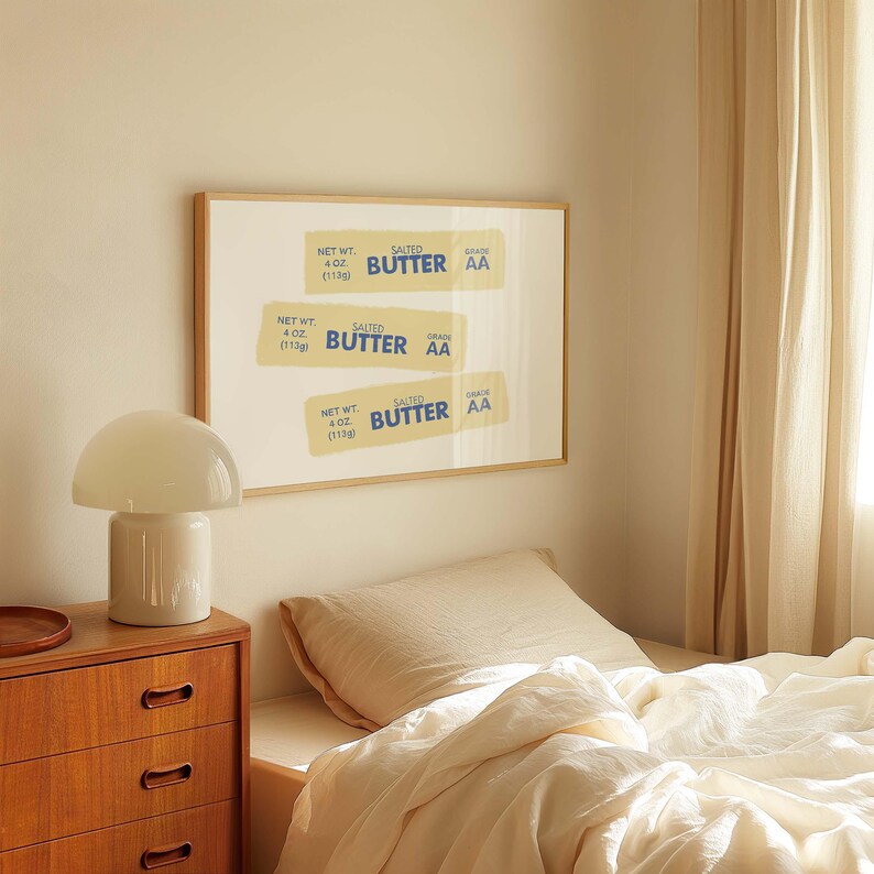 May include: A framed print of three vintage-style butter labels with the text "SALTED BUTTER GRADE AA" and the net weight of 4 oz (113g). The print is hanging on a wall in a bedroom with a white lamp and a bed with a beige linen duvet.