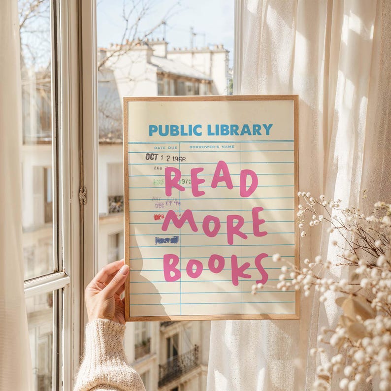 May include: A framed print resembling a vintage library card. The top text reads "PUBLIC LIBRARY" in blue, with "READ MORE BOOKS" in pink. The print is held near a window with a view of buildings and white curtains.