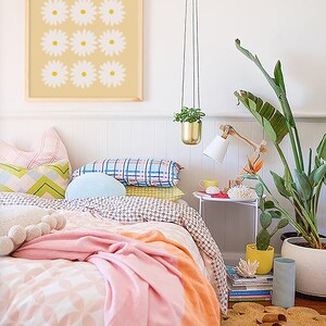May include: A bedroom with a white bed with a pink and orange blanket. There are several pillows on the bed, including a blue pillow. There is a framed print of white flowers on a yellow background hanging on the wall. There is a plant hanging from the ceiling and a plant in a white pot on the floor. There is a small table with a lamp and a stack of books next to the bed.