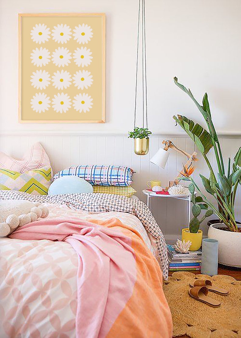 May include: A bedroom with a white bed with a pink and orange blanket. There are several pillows on the bed, including a blue pillow. There is a framed print of white flowers on a yellow background hanging on the wall. There is a plant hanging from the ceiling and a plant in a white pot on the floor. There is a small table with a lamp and a stack of books next to the bed.