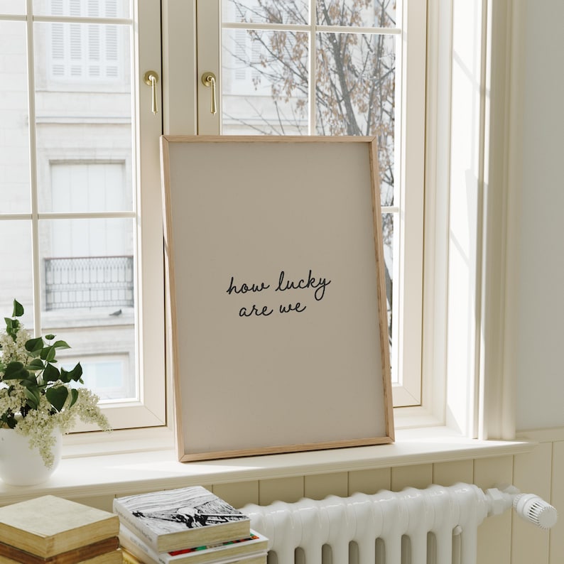 Puede incluir: Un estampado enmarcado con el texto "how lucky are we" en un estilo manuscrito. El estampado tiene un fondo beige claro y un marco de madera natural. Se exhibe en un alf&eacute;izar junto a una ventana.