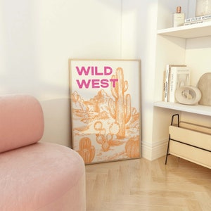 May include: A framed print with the words "WILD WEST" in pink lettering and a drawing of a desert landscape with a large cactus in the center.