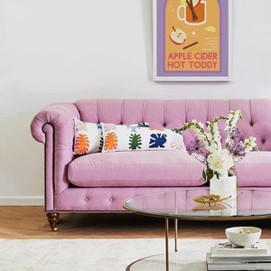 May include: A pink tufted sofa with two colorful pillows and a glass-top coffee table with a marble base. A vase of white and purple flowers sits on the table. A framed print with the text "Apple Cider Hot Toddy" hangs on the wall.