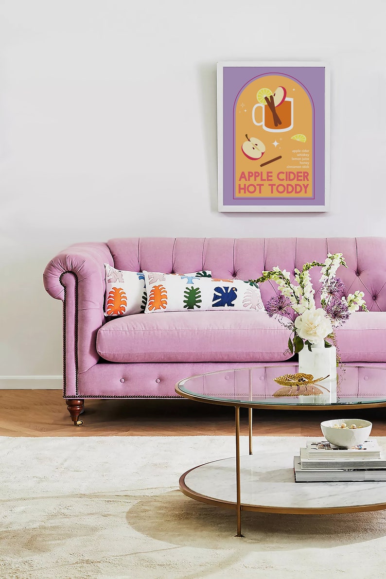 May include: A pink tufted sofa with two colorful pillows and a glass-top coffee table with a marble base. A vase of white and purple flowers sits on the table. A framed print with the text "Apple Cider Hot Toddy" hangs on the wall.
