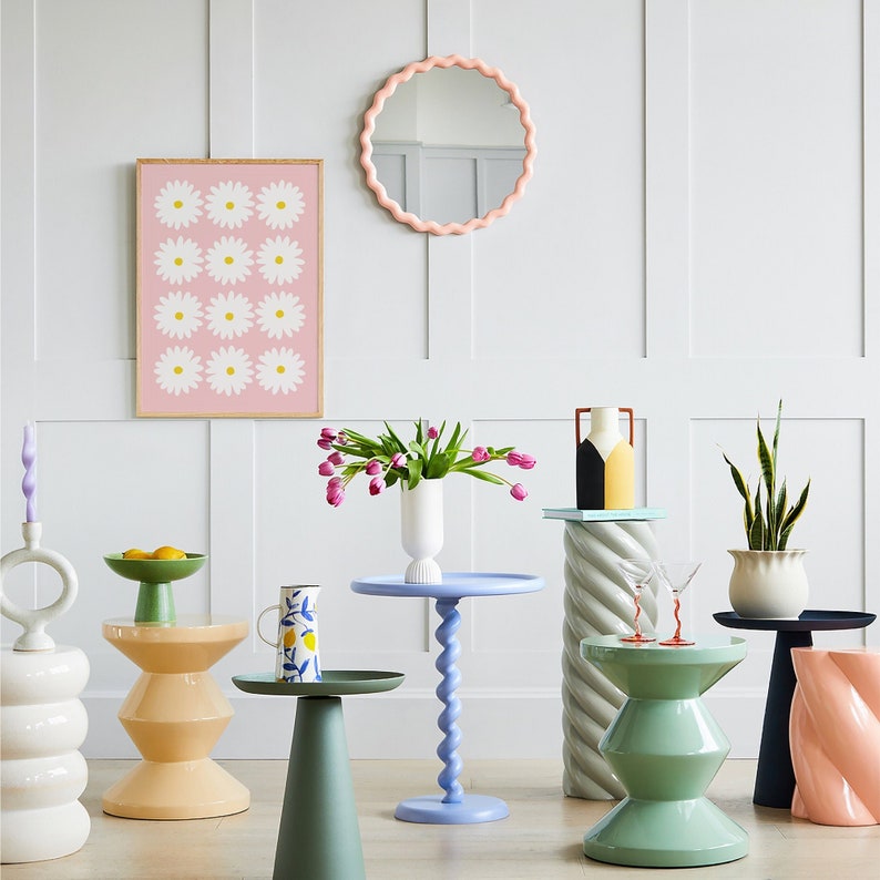 May include: A room with a pink and white daisy print framed artwork, a pink scalloped mirror, and several side tables with different colored bases. The tables are decorated with vases, plants, and glasses.