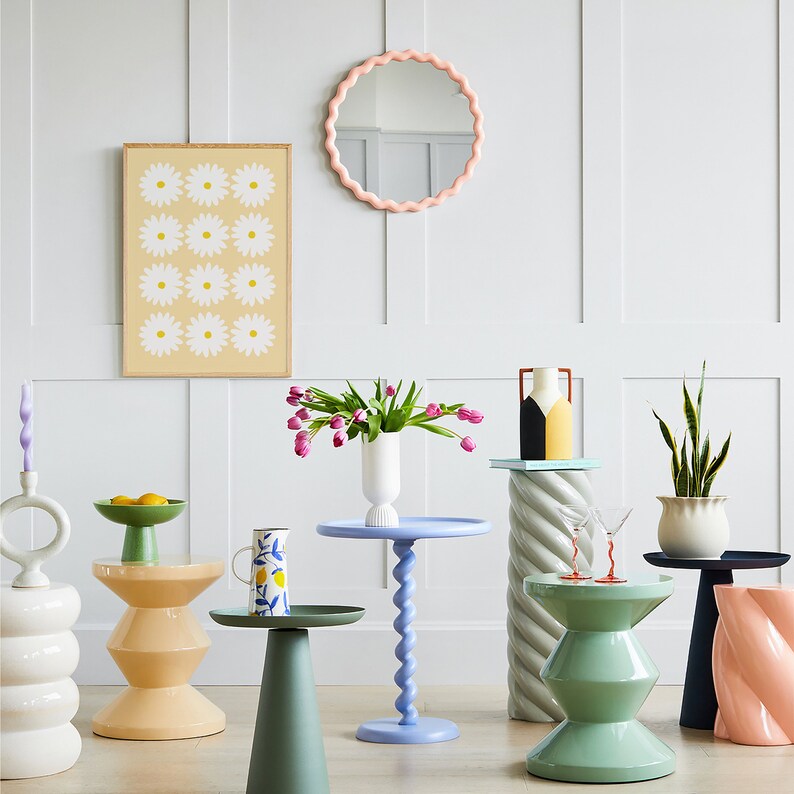 May include: A room interior with a white wall, a pink scalloped mirror, and several colourful side tables with different designs. The tables are decorated with flowers, a vase, a pitcher, and a bowl of fruit.