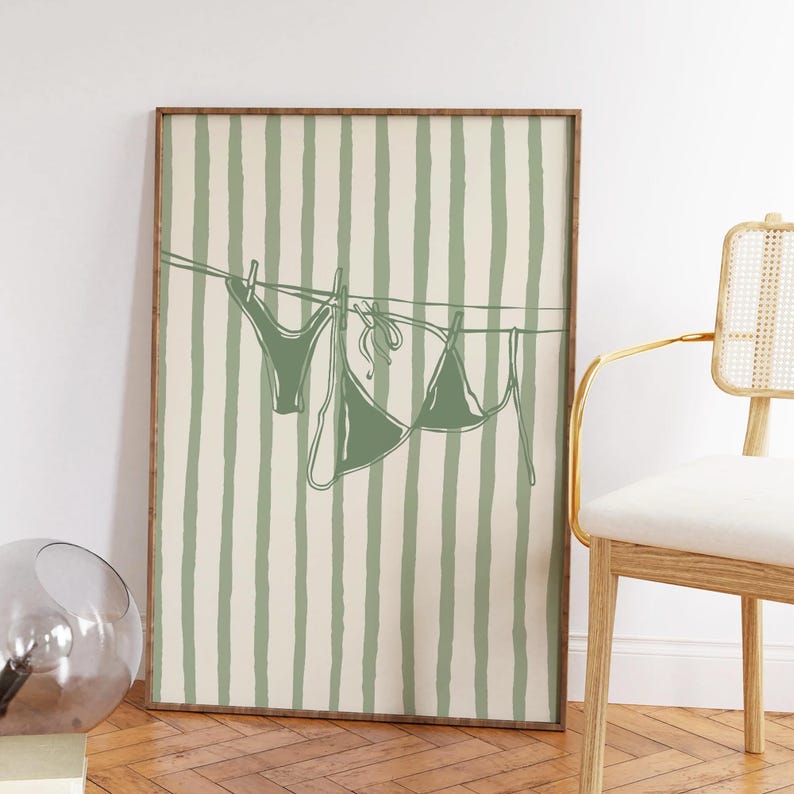 Puede incluir: Impresi&oacute;n art&iacute;stica enmarcada con una ilustraci&oacute;n minimalista de un conjunto de bikini verde colgado de un tendedero. El fondo tiene rayas verticales en verde claro y crema. La obra de arte se exhibe en una habitaci&oacute;n con una silla.
