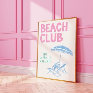Pu&ograve; includere: Una stampa incorniciata con il testo "Beach Club" in lettere rosa e "The ocean is calling" in lettere verdi. La stampa presenta un ombrello da spiaggia blu e due sedie a sdraio.