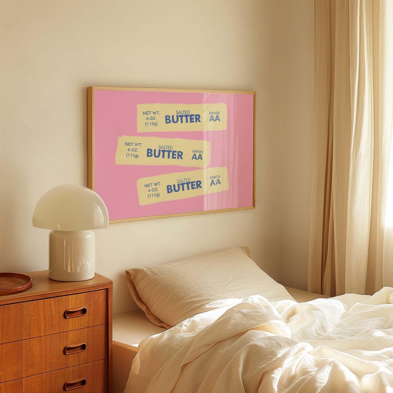 Peut inclure: Une affiche encadr&eacute;e repr&eacute;sentant trois paquets de beurre sur fond rose. Chaque paquet porte l'inscription "SALTED BUTTER" et "GRADE AA", avec un poids net de 113g. L'affiche est au-dessus d'un lit, avec une lampe blanche et une commode en bois.