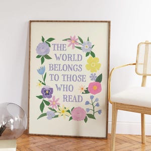 以下が含まれることがあります： 白い背景と花の縁取りのある額装されたプリント。"The world belongs to those who read"というテキストが紫色の文字で印刷されています。
