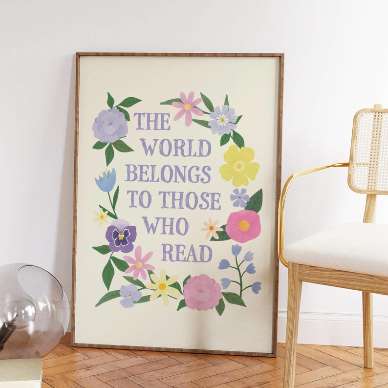 以下が含まれることがあります： 白い背景と花の縁取りのある額装されたプリント。"The world belongs to those who read"というテキストが紫色の文字で印刷されています。