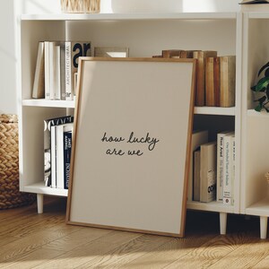 Puede incluir: Un cuadro enmarcado con el texto "how lucky are we" en un estilo manuscrito. El cuadro tiene un fondo beige claro y un marco de madera natural. El cuadro est&aacute; apoyado contra una estanter&iacute;a blanca llena de libros.