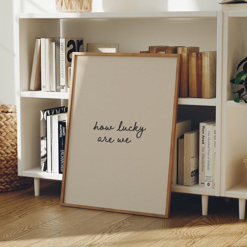 Puede incluir: Un cuadro enmarcado con el texto "how lucky are we" en un estilo manuscrito. El cuadro tiene un fondo beige claro y un marco de madera natural. El cuadro est&aacute; apoyado contra una estanter&iacute;a blanca llena de libros.