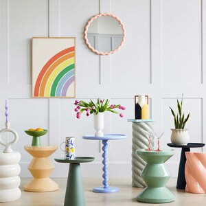 Peut inclure: Une pi&egrave;ce avec un mur blanc et un miroir rose &agrave; coquille. Il y a plusieurs tables d'appoint avec des plateaux et des pieds de diff&eacute;rentes couleurs. Une table a un plateau bleu et un pied en spirale. Une autre table a un plateau vert et un pied &agrave; motif g&eacute;om&eacute;trique. Une troisi&egrave;me table a un plateau jaune et un pied &agrave; motif g&eacute;om&eacute;trique. Il y a un vase avec des fleurs roses sur la table bleue. Il y a un pichet &agrave; motif floral sur la table verte. Il y a un bol avec des fruits sur la table jaune.