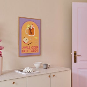 May include: A framed illustration of a mug of apple cider hot toddy with ingredients listed below. The illustration is in a muted orange, yellow, and purple color scheme. The frame is light brown wood.