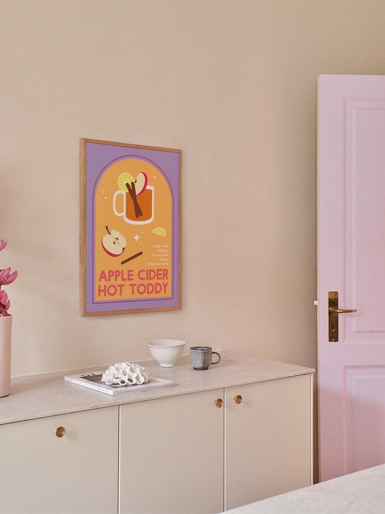 May include: A framed illustration of a mug of apple cider hot toddy with ingredients listed below. The illustration is in a muted orange, yellow, and purple color scheme. The frame is light brown wood.