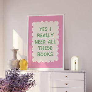 May include: A pink and white poster with the text "YES I REALLY NEED ALL THESE BOOKS" in green lettering. The poster is hanging on a white wall above a white dresser with a lamp and a vase of purple flowers.