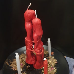 May include: Red figure candles tied together with red string, surrounded by white and red taper candles, herbs, and crystals on a black plate. The candles are in the shape of a man and woman.