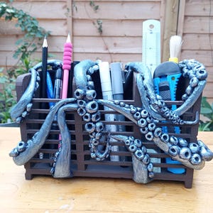 May include: A dark brown rectangular desk organizer featuring sculpted silver octopus tentacles wrapped around the exterior. The organizer holds various pens, pencils, and a utility knife, with a white ruler showing 8 inches in height.