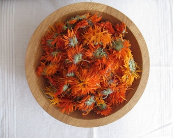 Può includere: Una ciotola rotonda in legno chiaro riempita con una collezione di capolini di calendula essiccati di colore arancione e giallo. I petali sono vivaci e strutturati, mostrando varie sfumature di arancione tramonto e oro, con piccoli calici verdi.
